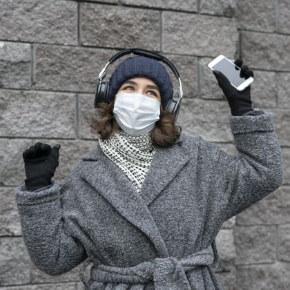 面罩戴着医用口罩的女人在城里用耳机和智能手机听音乐移动妇女女性