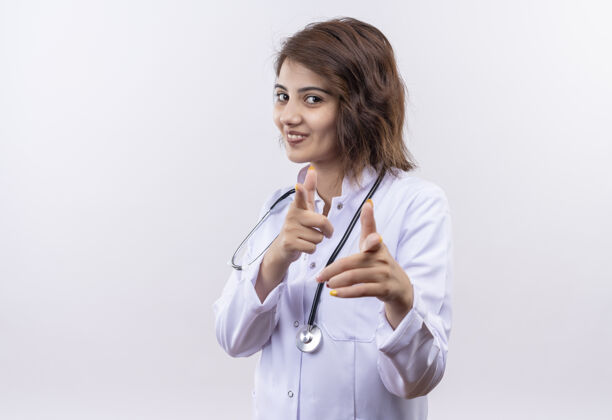 女人身穿白大褂 手持听诊器的年轻女医生微笑着 用食指指着白墙 双手高举医生听诊器站着