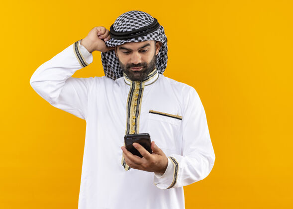 穿身着传统服装的阿拉伯商人站在橙色的墙上 看着智能手机的屏幕困惑不已困惑屏幕手机