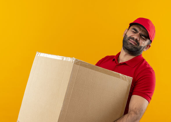 包装留着胡须的送货员穿着红色制服 戴着帽子 手里拿着一个大箱子 站在橘黄色的墙上 承受着沉重的重量拿着制服帽子