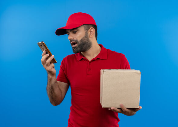站立有胡子的送货员穿着红色制服 戴着帽子 拿着一个盒子 站在蓝色的墙上用他的智能手机记录语音信息男手持使用
