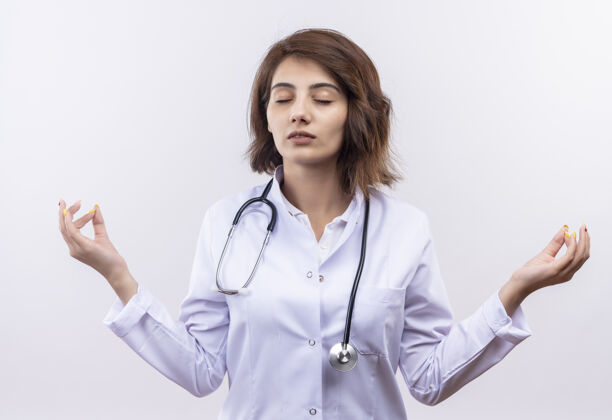 站着年轻的女医生 穿着白大褂 戴着听诊器 闭着眼睛放松 做着冥想的姿势 手指站在白墙上听诊器外套手势