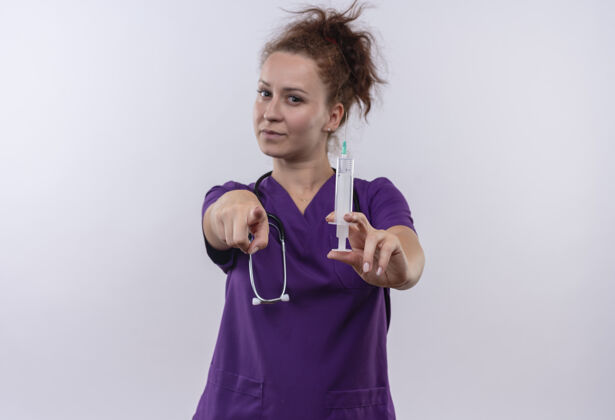 听诊器年轻的女医生 穿着医疗制服 手持听诊器 手指指着注射器 严肃的脸站在白色的墙上看穿脸