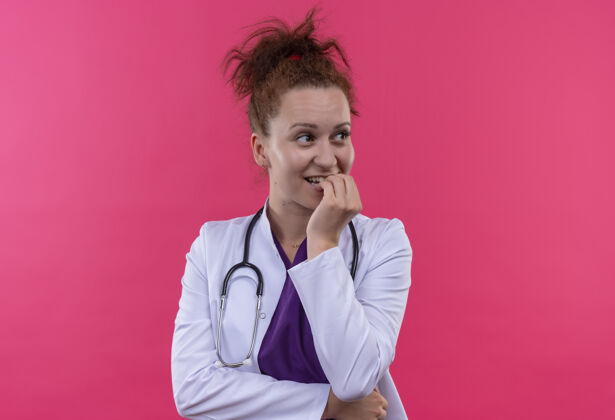 站年轻的女医生穿着白大褂 手持听诊器 站在粉红色的墙上 焦急而紧张地咬着指甲看听诊器担心