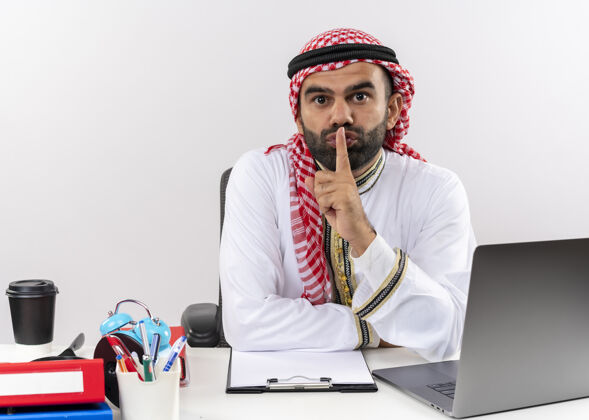 电脑身着传统服装的阿拉伯商人坐在桌旁 拿着笔记本电脑 手指放在嘴唇上做着沉默的手势 在办公室工作阿拉伯语手指笔记本电脑