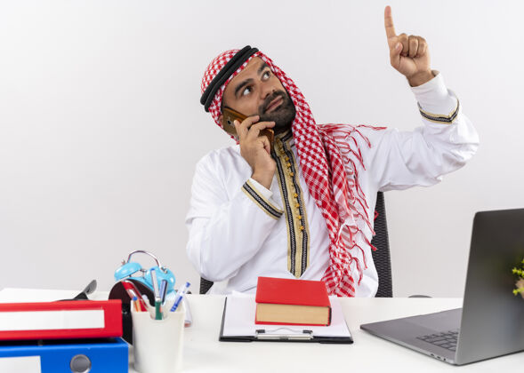 传统身着传统服装的阿拉伯商人坐在桌子旁用手机指着办公室工作交谈商人穿