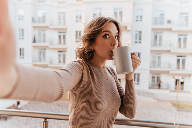 寒冷穿着棕色毛衣的有趣女孩在阳台上喝咖啡迷人的年轻女子用一杯茶自拍年轻卷发公寓