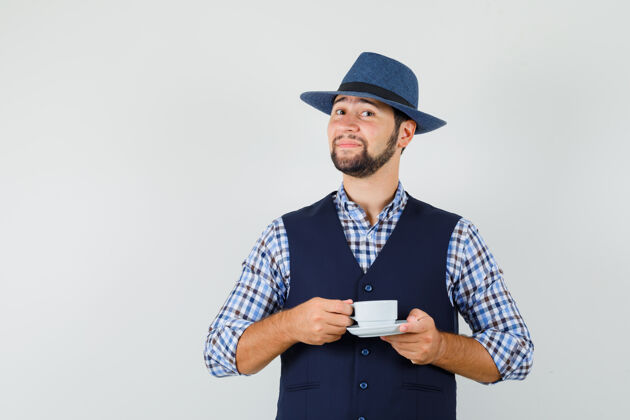 男人年轻人拿着杯子和茶托 穿着衬衫 背心 戴着帽子 看上去很自信 正对着窗外发型肖像聪明