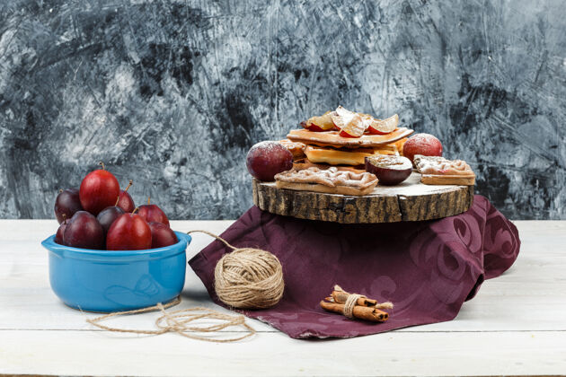 甜点把华夫饼和饼干放在圆形柳条垫子上 放上一碗李子 勃艮第桌布 在深蓝色大理石和白色木板表面画上提示水平食物饼干糖