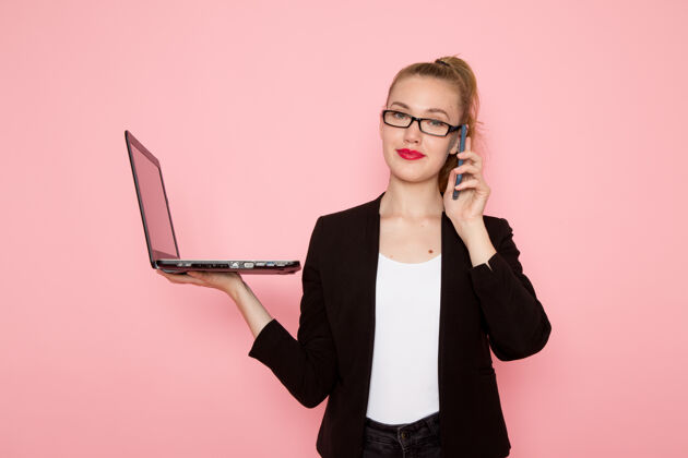 女性上班族身穿黑色夹克的女上班族在粉色墙上打电话和使用笔记本电脑的正面图工人商务女性前台