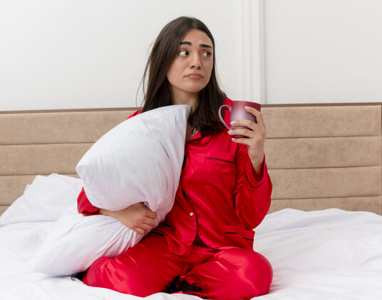 卧室穿着红色睡衣的年轻漂亮女人坐在床上 拿着一个枕头 拿着一个杯子 一边看一边 在卧室的室内灯光背景下感到困惑抱着红色枕头