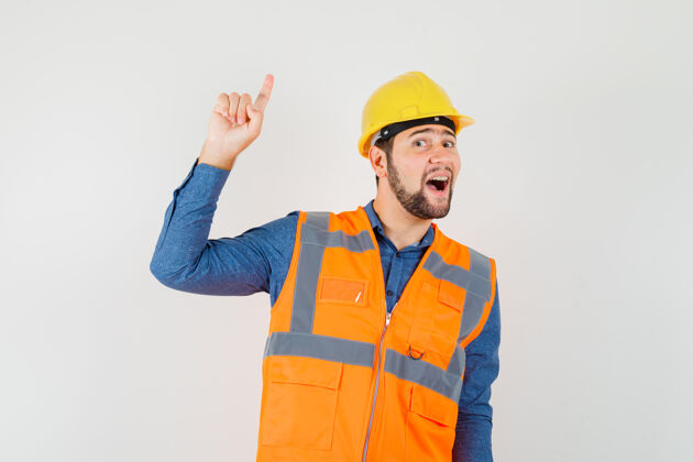 衬衫年轻的建筑工人指着上面的衬衫 背心 头盔 看起来很快乐前视图工具指点建筑