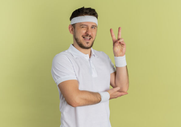 运动员身穿白衬衫 头箍的年轻健身男子面带微笑 眨眼 站在橄榄墙上展示胜利标志年轻看健康