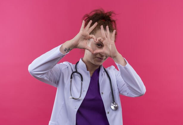 医生年轻的女医生身穿白色外套 手持听诊器 站在粉色的墙上 用手指做着心脏的手势 微笑着看着手指站手势女人