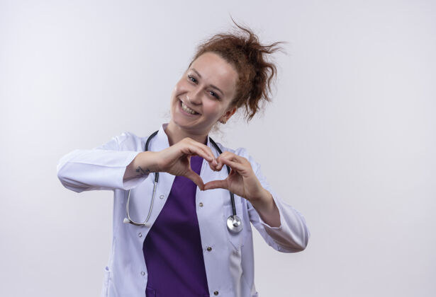 站立年轻的女医生穿着白大褂 手持听诊器做心脏手势 手指放在胸前 微笑着积极快乐地站在白墙上佩戴胸部女人