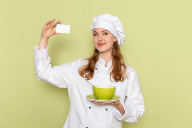 烹饪身着白色厨师服的女厨师的正面图 绿色墙上有名片和盘子人家庭主妇食物