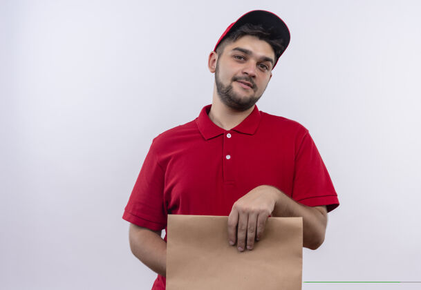 微笑年轻的送货员穿着红色制服 戴着帽子 手里拿着纸包 微笑着自信制服年轻纸