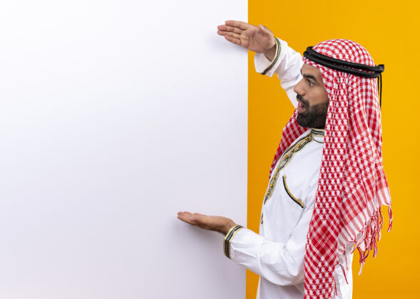手臂身着传统服装的阿拉伯商人站在橙色的墙上 指着空白的广告牌 手臂看起来很惊讶指向传统空白