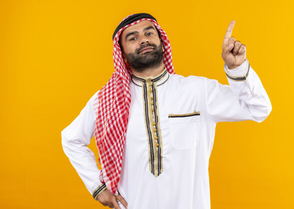 穿身着传统服装的阿拉伯商人自信地用手指着站在橙色墙上的一边商务人士指手指