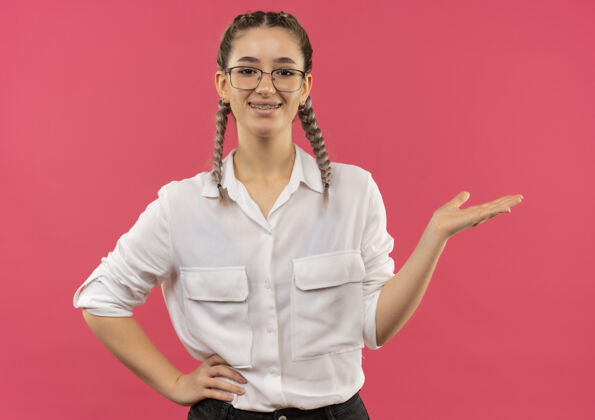 衬衫戴着眼镜 梳着辫子 穿着白衬衫的年轻女学生 用手举着什么东西 面带微笑地站在粉红色的墙上 望着前方手年轻人眼镜
