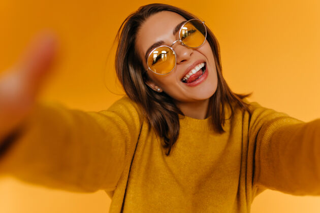 明亮幸福的棕褐色毛衣女人自拍和笑戴墨镜的温文尔雅女孩在橘色的墙上给自己拍照放松奇迹肖像
