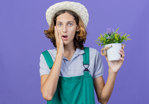 男人年轻的园丁穿着连体衣戴着帽子拿着盆栽植物感到惊讶连身衣植物抱着