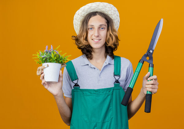 持有年轻的园丁 穿着连体衣 戴着帽子 拿着树篱剪和盆栽植物花园男人微笑