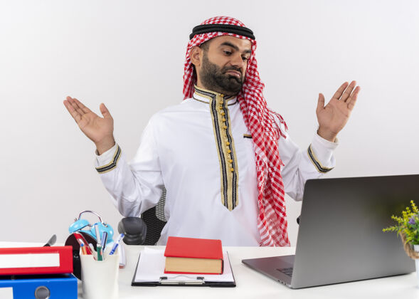 桌子身着传统服装的阿拉伯商人坐在桌子旁 手提电脑看起来很困惑 不知所措地举起双臂 在办公室工作时没有回答看笔记本电脑不知所措