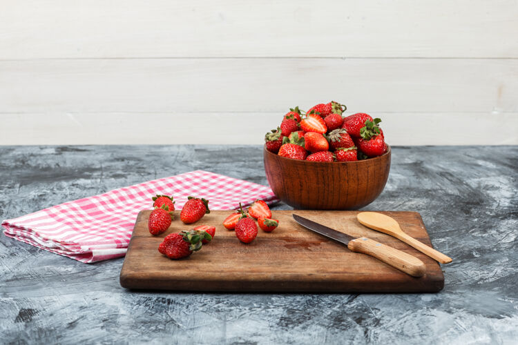烹饪用红色方格桌布和深蓝色大理石和白色木质背景上的一碗草莓 在木制砧板上关闭草莓和厨房用具水平有机糕点木板