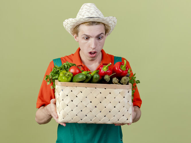 惊喜年轻的园丁穿着连体衣 戴着帽子 手里拿着装满蔬菜的箱子 站在灯光的背景下 看上去既惊讶又惊讶抱着连身衣年轻人