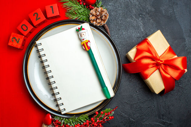 圣诞新年背景俯视图 餐盘上有螺旋笔记本装饰配件 红色餐巾上有杉木树枝和数字 黑色桌子上有红丝带礼物餐盘笔记本餐巾