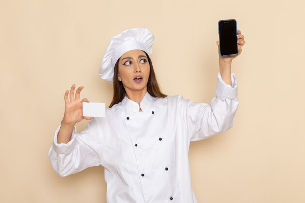 厨房身穿白色厨师服的年轻女厨师正拿着智能手机和卡片站在浅白的墙上烹饪移动女性