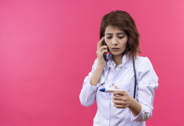 外套穿着白大褂的年轻女医生拿着听诊器拿着数字体温计一边担心地看着它一边讲手机站着说话医生