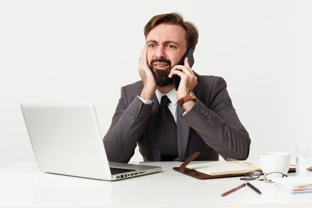 办公室困惑的年轻黑发男子胡子穿着正式的衣服 而工作在白色的墙壁上 头靠在举起的手上 困惑地看着他的笔记本电脑屏幕衣服衬衫男性