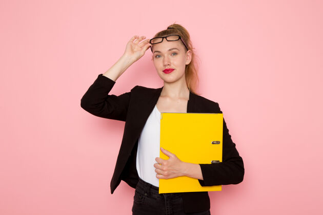 严格身穿黑色紧身夹克的女上班族正拿着黄色文件站在浅粉色的墙上夹克工人女