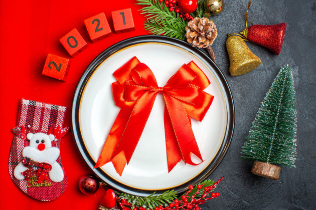 上衣新年背景俯视图 餐盘上有红丝带装饰配件杉木树枝和数字圣诞袜放在黑色桌子上圣诞树旁的红色餐巾上树枝风景袜子