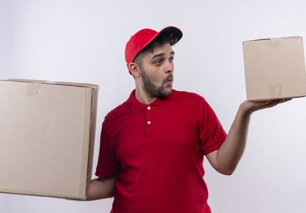 做年轻的送货员穿着红色制服 戴着帽子 手里拿着大纸箱 看上去很困惑 试图做出一个选择拼图纸板盒子