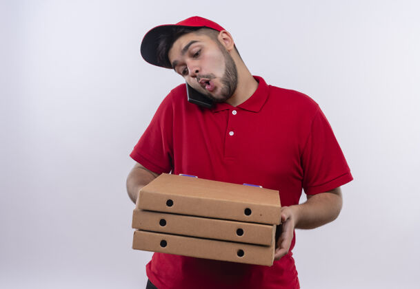 男人年轻的送货员穿着红色制服 戴着帽子 手里拿着披萨盒 忙着打手机帽子拿着非常