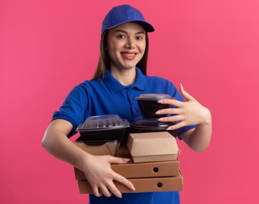 漂亮穿着制服的微笑漂亮的女送货员拿着比萨饼盒上的食品包和容器 这些比萨饼盒被隔离在粉红色的墙上 并留有复印空间微笑披萨持有