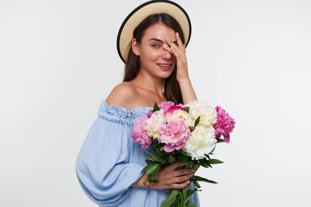 长发十几岁的女孩微笑着 深色长发的女人戴着帽子 穿着蓝色漂亮的裙子拿着一束花 隔着白色的墙隔着手指看着手指年轻面部