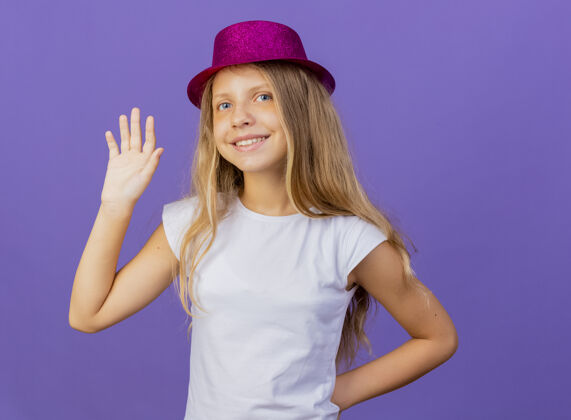 漂亮戴着节日礼帽的漂亮小女孩挥舞着快乐而积极的手 紫色背景下站着生日派对的概念生日节日帽子