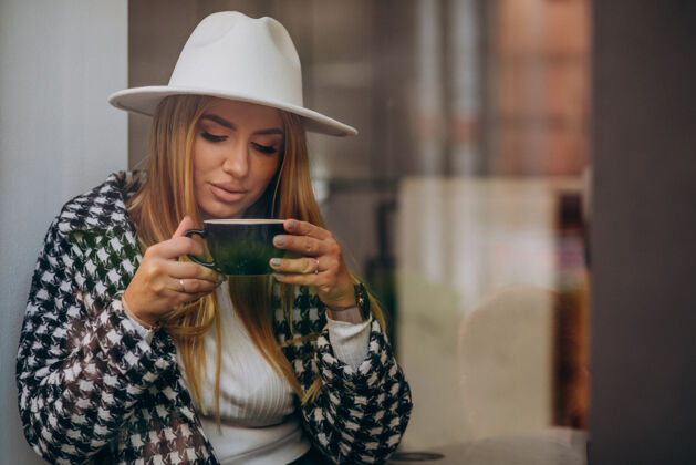 坐着在咖啡馆喝咖啡的女人 坐在玻璃后面看帽子思考