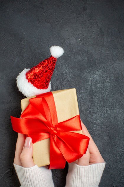 蝴蝶结手持精美礼物的垂直视图 黑色背景下的圣诞老人帽子旁边有蝴蝶结状的缎带漂亮的礼物帽子长袜