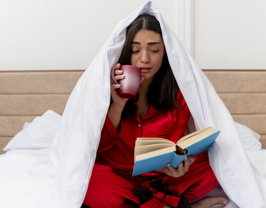 书穿着红色睡衣的年轻漂亮女人坐在床上 用毯子裹着一杯咖啡 在卧室的室内灯光背景下看书睡衣红色室内