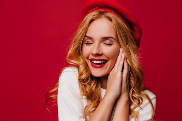 年轻穿着法国贝雷帽的可爱白人女性表达灵感温文尔雅的金发女孩闭着眼睛笑着站在红墙上表情室内情绪