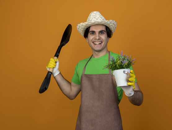 人年轻的男园丁面带微笑 身着制服 戴着园艺帽 戴着手套 手持铁锹 花盆里的花被隔离在橙色的墙上帽子人花