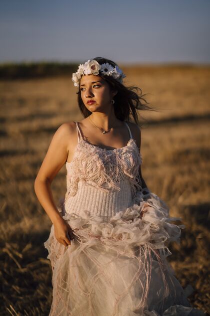 乡村一个年轻的白人女性穿着白色的裙子和白色的花环摆在一个领域的垂直镜头农业和平日落