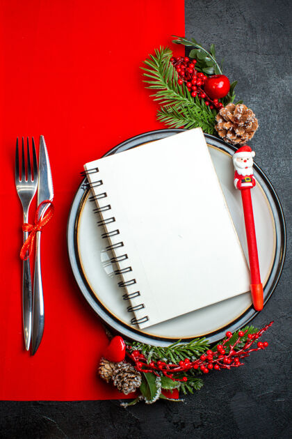 餐巾一个螺旋笔记本的垂直视图和一支钢笔放在餐盘上 装饰配件杉木树枝和餐具放在红色餐巾上冷杉餐具冬青