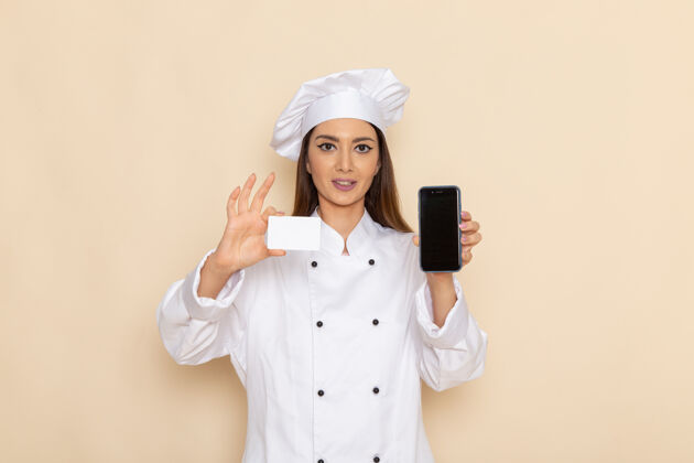 食物身穿白色厨师服的年轻女厨师的正视图 在浅白的墙上拿着电话和名片电话专业制服