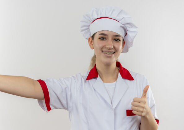 拇指面带微笑的年轻漂亮厨师身着厨师制服 戴着牙套 张开的手臂在空白处向上伸出大拇指漂亮向上年轻